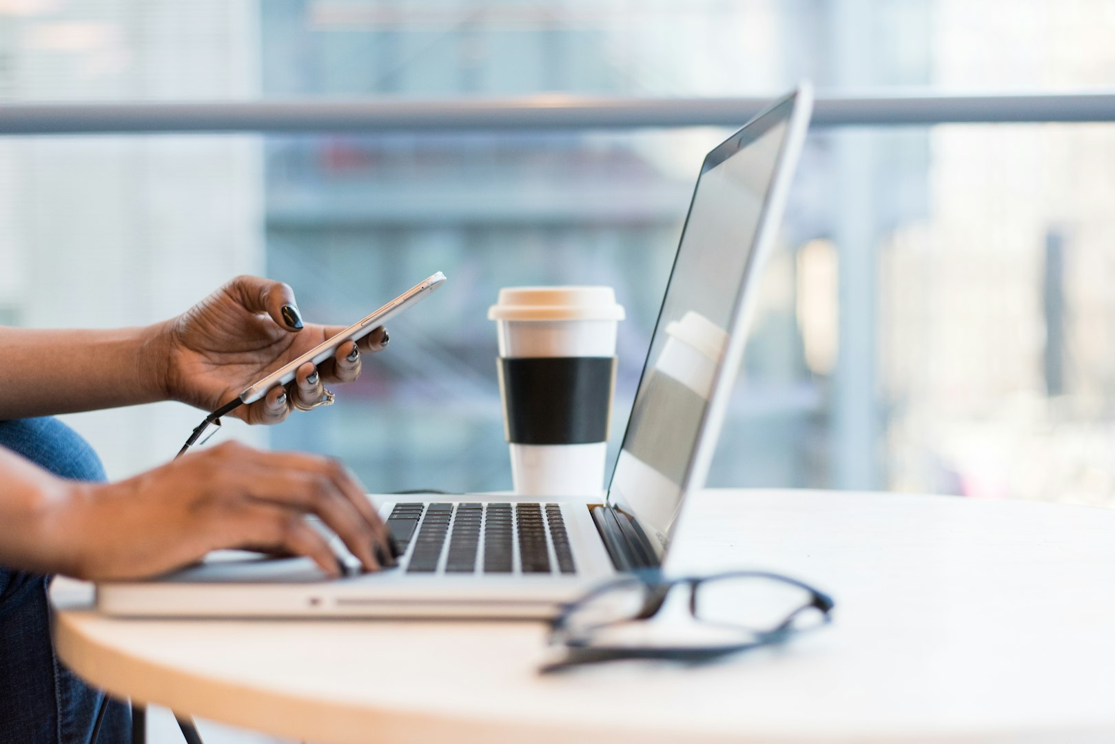 Person arbetar effektivt vid ett skrivbord med en laptop och en smartphone, en kopp kaffe bredvid sig, med ett ljust fönster i bakgrunden.